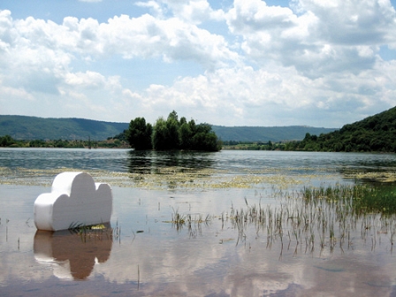 Eine aus Styropor ausgeschnittene Wolke schwimmt auf einem Teich, am Himmel stehen Wolken